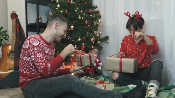 La familia feliz está celebrando la Navidad juntos en casa, abriendo regalos de caja de regalo envueltos en lazo rojo. Pareja joven sentada cerca del árbol de Navidad con regalos y disfrutando de las vacaciones. Espíritu de Navidad — Vídeo de stock