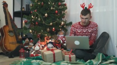 Noel kırmızı kazaklı yakışıklı genç adam Noel ağacının yanında bir sürü hediyeyle oturuyor, Noel 'de ya da yeni yıl gecesinde dizüstü bilgisayarda çalışıyor. Noel ruhu ve kutlama.