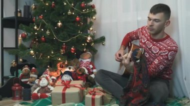 Genç adam akustik gitar çalıyor Noel ağacının yanında oturuyor ve şarkılar söylüyor, 2021 yılını evde tek başına kutluyor. Noel ruhu, tatil ve kutlama konsepti