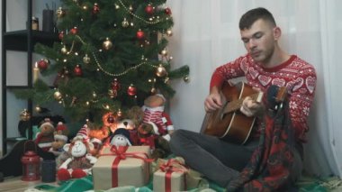 Kırmızı kış kazağı giyen yakışıklı adam Noel ağacının yanında ekoseli oturuyor ve gitar çalıyor, evde yalnız başına yeni yılı kutluyor. Noel ruhu, yeni yıl 2021. Kutlama ve eğlence konsepti