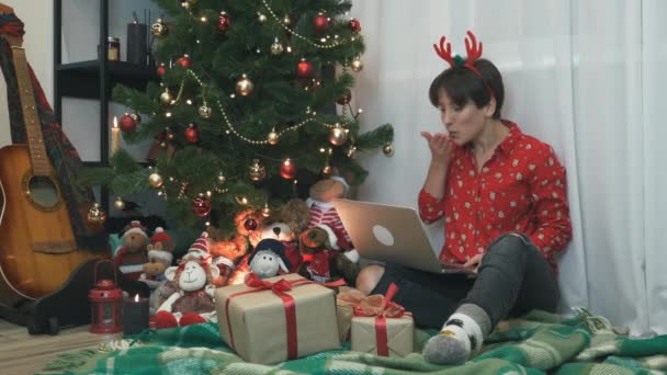 Mujer joven festiva en ropa de fiesta de Navidad está sentado cerca del árbol de Navidad, celebrando el Año Nuevo solo en casa, haciendo una llamada de conferencia con amigos y familiares, enviando besos aéreos y riendo — Vídeo de stock