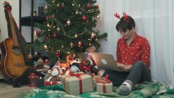 Charmante jeune femme festive travaille sur ordinateur portable à Noël ou au Nouvel An soir. Happy femelle est assis près de l'arbre de Noël avec beaucoup de cadeaux, en utilisant un carnet, bavarder avec des amis — Video
