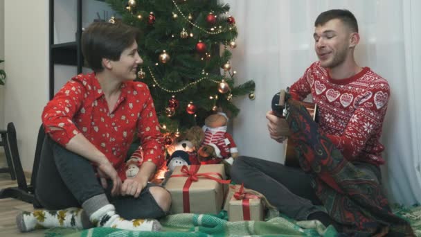 คู่รักหนุ่มสาวที่ตื่นเต้นในเสื้อผ้าคริสต์มาสฤดูหนาว ฉลองปีใหม่และคริสต์มาสด้วยกันที่บ้าน ผู้ชายกําลังเล่นกีตาร์และร้องเพลงคริสต์มาสในวันหยุดฤดูหนาว ปรบมือและมีความสุข — วีดีโอสต็อก