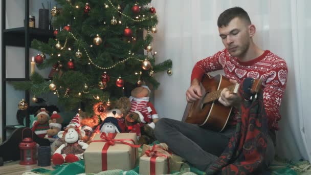 Hombre guapo en suéter rojo de invierno está sentado en cuadros cerca del árbol de Navidad y tocando la guitarra, celebrando la noche de Año Nuevo solo en casa. Espíritu de Navidad, Año Nuevo 2021. Celebración y concepto divertido — Vídeo de stock