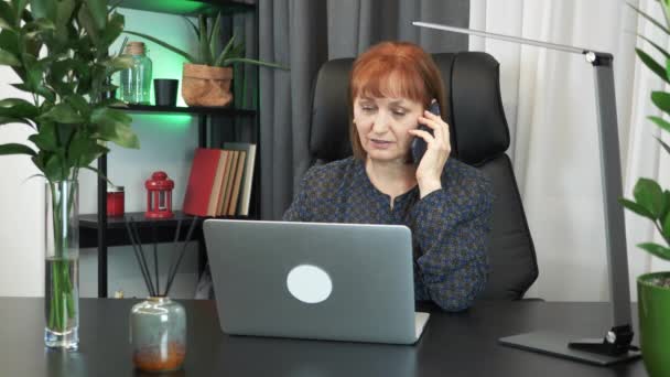 Úspěšná podnikatelka mluví na chytrém telefonu s partnery, zatímco sedí na svém moderním pracovišti v kanceláři. Soustředěná žena pracuje na notebooku a mluví po telefonu na kancelářském stole — Stock video
