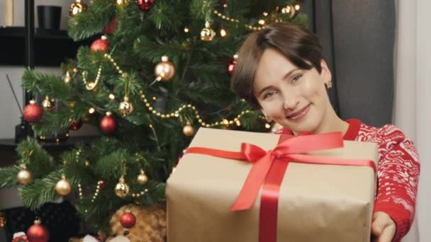 Mulher está dando presente de Natal embrulhado em papel artesanal com arco vermelho. Mulher sorridente está segurando caixa de presente de Ano Novo. Celebrando Feliz Natal ou Feliz Ano Novo 2021 — Vídeo de Stock