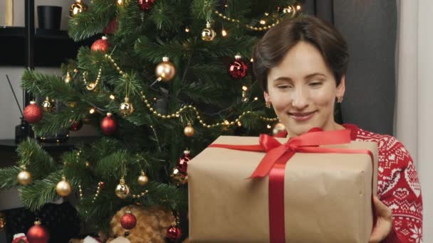 Mujer feliz joven está sacudiendo la caja de regalo con regalo de Navidad sentado cerca del árbol de Navidad. Mujer sostiene regalo de Navidad envuelto con cinta roja. Celebración de Año Nuevo y Nochebuena — Vídeo de stock