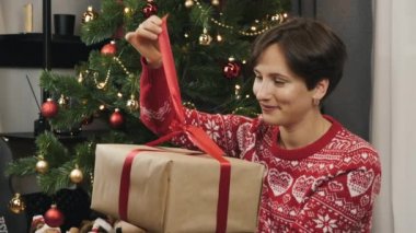 Kadın, yeni yıl ağacının altında oturarak büyük bir Noel hediyesi paketi açıyor. Mutlu gülümseyen kadın Noel hediyesini açıyor ve kırmızı kurdeleyi çözüyor. Mutlu yıllar 2021