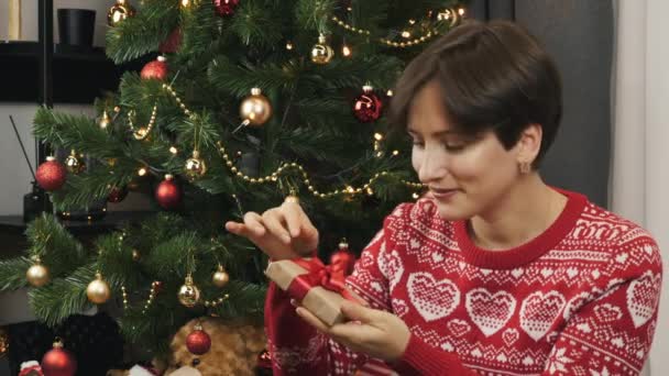 Mulher está sentada perto da árvore de Natal e segurando pequena caixa de presente para o Ano Novo. Feliz Natal e Ano Novo Concept celebração. Mulher detém presente em forma de caixa de presente em suas mãos e sorrisos — Vídeo de Stock