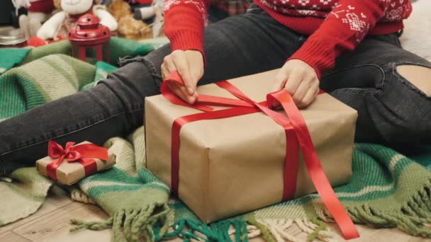 Manos de mujer atando cinta roja alrededor de la caja de regalo de Navidad. Caja de regalo de Navidad o Año Nuevo cerca del árbol de Año Nuevo. La hembra está envolviendo un regalo festivo sorpresa. Preparación para la temporada de invierno de vacaciones — Vídeo de stock