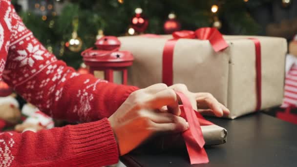 As mãos femininas finalizam o presente de Natal envolto em papel craftool na mesa de madeira. Mulher está embrulhando caixa de presente, enfaixando fita vermelha e amarrado em arco. Preparando-se para feriados de Ano Novo e Natal — Vídeo de Stock