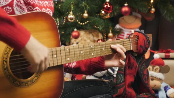 Woman speelt kerstliedje op akoestische gitaar zittend op de vloer in de buurt van versierde nieuwjaarsboom in slinger lichten. Gelukkige familie viert kerstavond. Vrolijk kerstconcept — Stockvideo