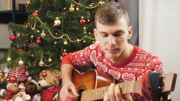 L'uomo sorridente in maglione festivo rosso sta suonando la chitarra e cantando canzoni natalizie seduto vicino all'albero decorato di Capodanno a casa. Celebrazione di Capodanno 2021 e Buon Natale — Video Stock