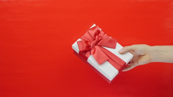 Hand visar vit låda med rött band rosett på röd bakgrund. Honan håller alla hjärtans dag gåva i handen. Alla hjärtans dag, internationella kvinnodagen — Stockvideo