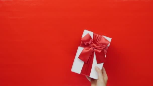 休日プレゼント。赤いリボン弓とバレンタインデーのギフトボックス。ロマンチックな挨拶だ。手は赤い背景に赤い弓で白い箱を保持しています。女性は手に誕生日プレゼントを示しています — ストック動画