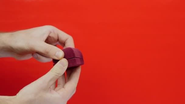 Mann hält Ehering in Box für Heiratsantrag auf rotem Hintergrund. Hand hält rote Schachtel mit Ehering. Geschenk zum Valentinstag — Stockvideo