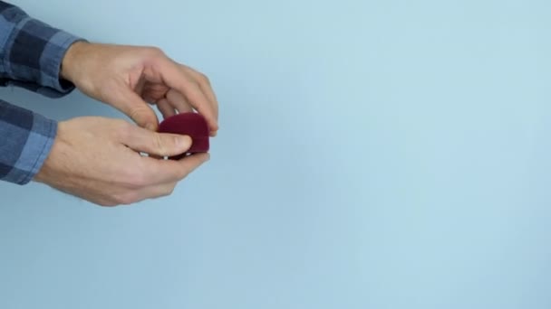 Hände öffnen ein rotes Kästchen mit Verlobungsring für einen Heiratsantrag auf blauem Hintergrund. Mann macht Heiratsantrag mit Ehering aus nächster Nähe — Stockvideo