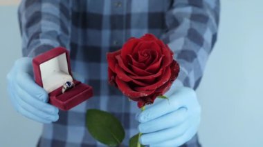 Mavi koruyucu tıp eldivenli adam evlilik yüzüğü ve kırmızı gülle evlenme teklif ediyor. Salgın karantina hijyeni. Tıbbi lastik eldivenli eller nişan yüzüklü küçük kırmızı kutuyu tutuyor.