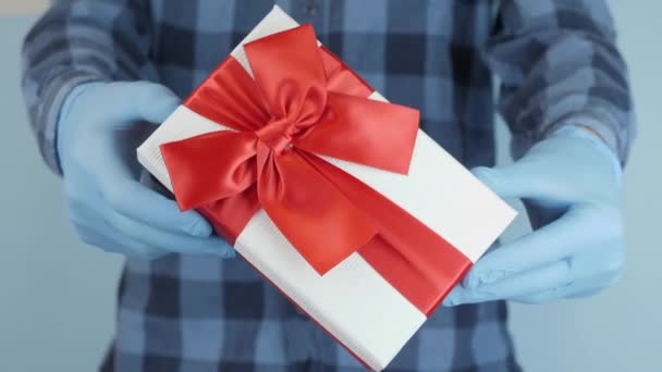 День святого Валентина і святкування свята з захистом від коронавірусу. Чоловічі руки в медичних гумових рукавицях показують подарунковий ящик з червоною стрічкою. Людина тримає подарункову коробку з червоним луком. — стокове відео
