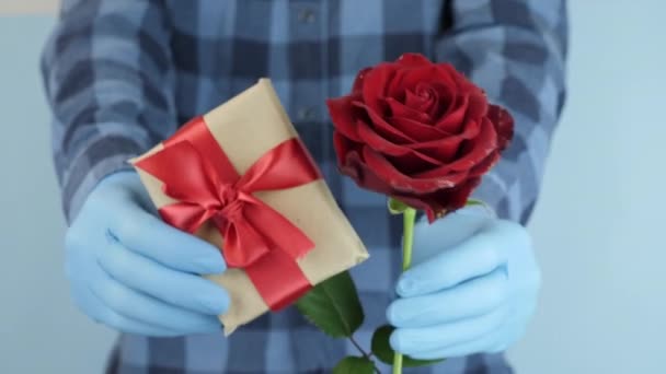 Male hands wearing medical rubber gloves are showing gift box present  and red rose, close up. Holiday celebration with protection from coronavirus covid19. Man in blue medical gloves gives gift — Stock Video