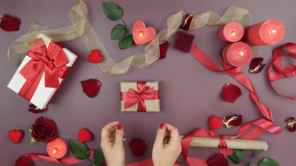 Hands are unwrapping present gift box with red ribbon bow on table. Festive background with roses, burning candles and gifts on pink or red background. Valentine's day gift concept — Wideo stockowe