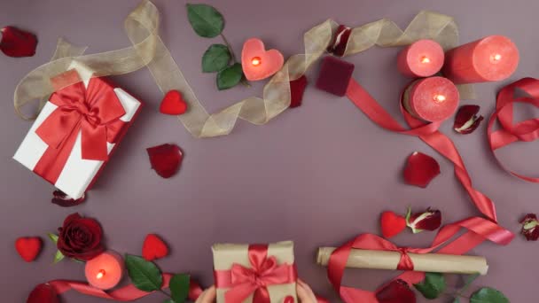 Women's hands put wrapped gift box with red ribbon bow on festive table with roses, burning candles and gifts on pink or red background. The concept of Valentine's day — Stock video