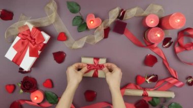 Female hands are tying red ribbon bow on wrapped gift box for St. Valentine's Day. Woman is preparing present gift for 14th of February. Table with roses, burning candles and gifts
