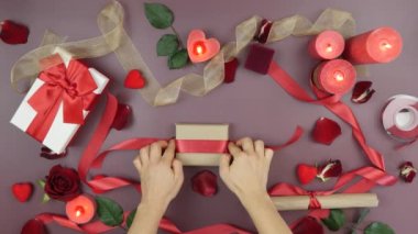 Kadınlar Sevgililer Günü için hediye kutusuna kırmızı kurdele bağlıyor. Arka planda güller, mumlar, hediyeler ve şöminelerle. Kadın Sevgililer Günü için hediye kutusu dekore ediyor.