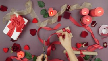 Bayanların elleri, neşeli Sevgililer Günü arka planında kırmızı kurdeleyi makasla kesiyor. Güller, mumlar, hediyeler ve şöminelerle. Kadın Sevgililer Günü için hediye kutusu dekore ediyor.