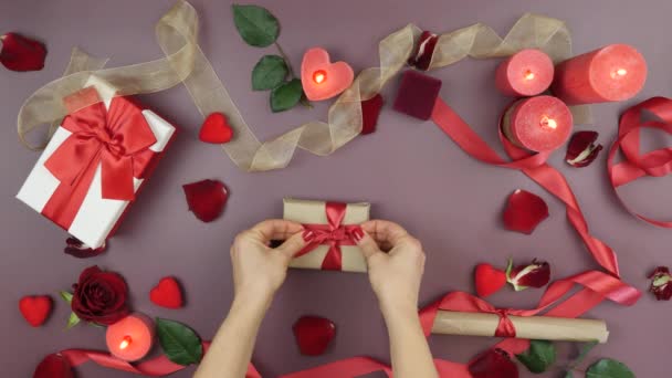 Female hands are tying red ribbon bow on wrapped gift box for St. Valentine's Day. Woman is preparing present gift for 14th of February. Table with roses, burning candles and gifts — Stockvideo