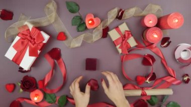 Hands are opening little red box with engagement ring for St. Valentine's Day on festive table background with roses, candles, gifts and hearths. Concept of love and romance