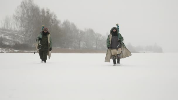 釣り棒と氷のドリルを持つ漁師は冬の釣りで凍った湖を通っています — ストック動画