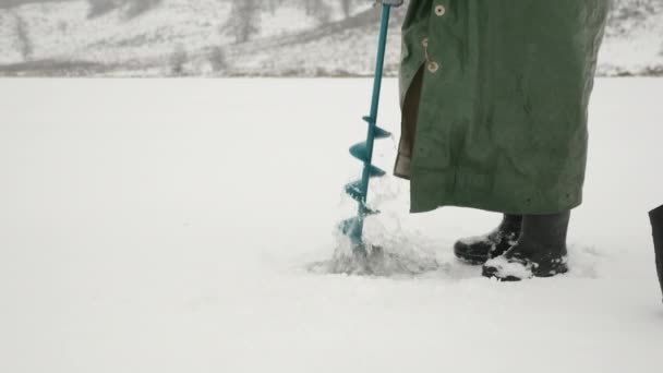 Rybář je vyvrtaná ledová díra na zamrzlém jezeře na zimním rybolovu. Muž s ledovým vrtákem v rukou — Stock video