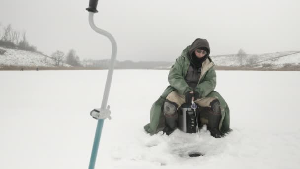 漁師は氷の穴と釣りの近くに座っている。男は湖で魚を捕っている — ストック動画