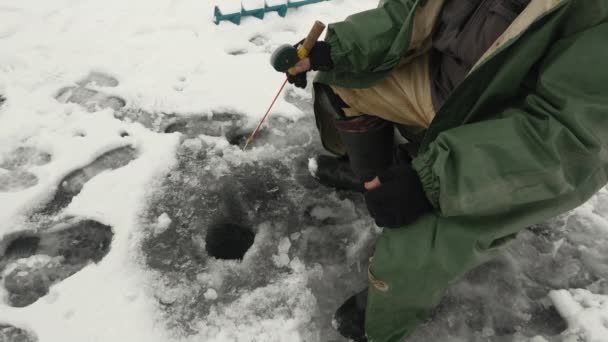 Ο άνθρωπος ψαρεύει στην τρύπα του πάγου. Ο ψαράς με καλάμι ψαρεύει ψάρια στο ποτάμι. — Αρχείο Βίντεο