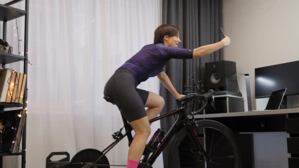 Feminino está pedalando no treinador de bicicleta inteligente em casa, tirando selfies no smartphone — Vídeo de Stock