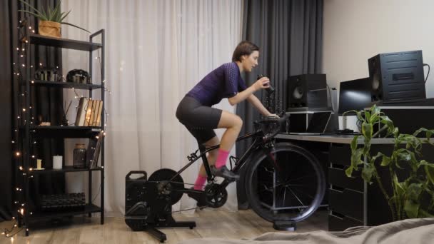 Γυναίκα εκπαιδεύεται σε σταθερό ποδήλατο, λαμβάνοντας μέρος σε online ποδηλατικούς αγώνες — Αρχείο Βίντεο