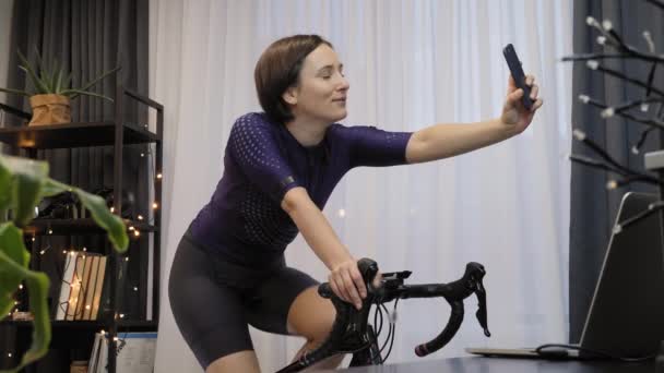 Mulher sorridente está tomando selfie no smartphone durante o treinamento em bicicleta estacionária — Vídeo de Stock