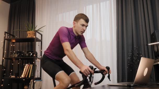 Joven atleta masculino está entrenando en bicicleta estacionaria en casa. Ciclismo interior — Vídeos de Stock