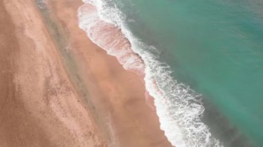 Kumlu sahil şeridinde deniz dalgaları kırılıyor. Derin mavi okyanus suyu ve beyaz köpüklü altın plaj