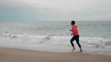 Kadın dalgaların üzerinde yalınayak koşuyor. Kadın, kumlu sahilde büyük okyanus dalgalarıyla koşuyor.