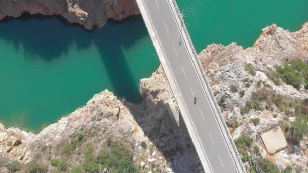 人は澄んだターコイズブルーの水と山の崖と川の上の橋の上に自転車に乗る — ストック動画