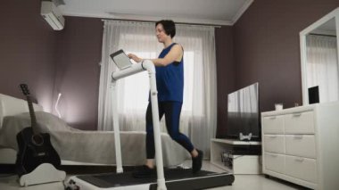 Kadın koşu bandında egzersiz yapıyor, tablette ekrana dokunuyor, çevrimiçi fitness antrenmanları izliyor.