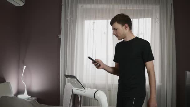 Mann trainiert zu Hause auf Laufband, Smartphone in der Hand. Laufen auf Laufband — Stockvideo
