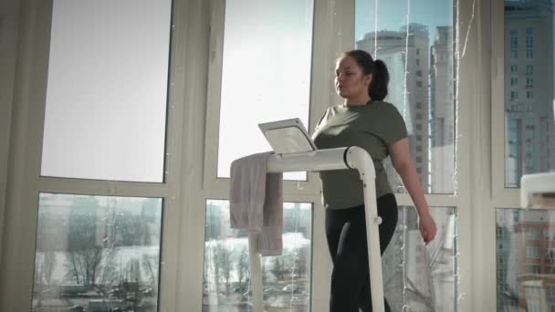 Mujer está entrenando en la cinta de correr, tocando la pantalla de la tableta, viendo noticias en Internet — Vídeos de Stock