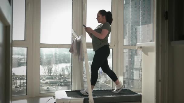 Frau trainiert auf Laufband, hält Smartphone in der Hand, tippt Nachrichten — Stockvideo