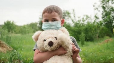Küçük sevimli çocuk elinde ayı oyuncağı, koruyucu tıbbi maske takıyor.