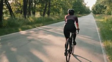 Gün batımında bisiklet sürerken, güneş ışınları yeşil ağaçların yapraklarını kırıyor. Gün doğumunda bisiklet süren kadın