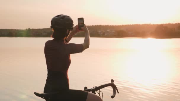 Kobieta rowerzystka stojąca na skraju wybrzeża, patrząca na zachód słońca i robiąca zdjęcia na smartfonie — Wideo stockowe