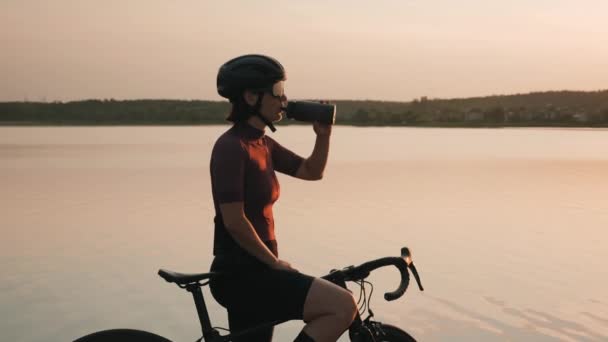 Велосипедист пьет воду на закате. Велосипедистка пьет воду после тяжелой тренировки — стоковое видео